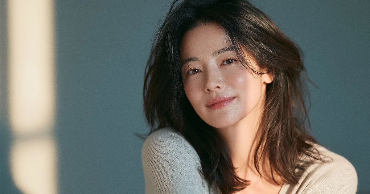 창가 빛이 비치는 푸른 회색 벽 앞에서 옅게 미소 지으며 고개를 살짝 기울인 여성의 얼굴