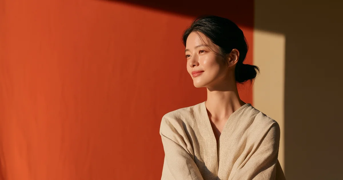 테라코타색 벽 앞에서 머리를 낮게 묶고 창가 빛을 받으며 옅게 미소 짓는 여성의 반측면