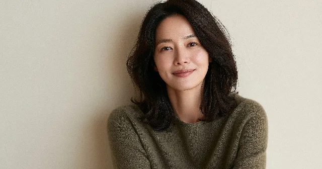 어깨까지 오는 머리를 풀고 올리브색 니트를 입은 40대 여성이 밝은 벽 앞에서 옆을 보며 웃는 모습