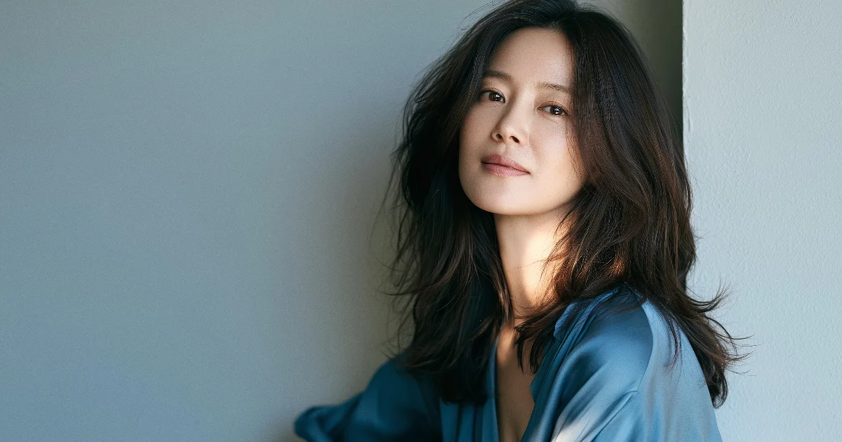 옅은 청회색 벽 앞에서 어깨 아래로 내려오는 웨이브 머리에 하늘색 실크 블라우스를 입은 여성이 창가 빛을 받으며 옅게 미소 짓는 반측면
